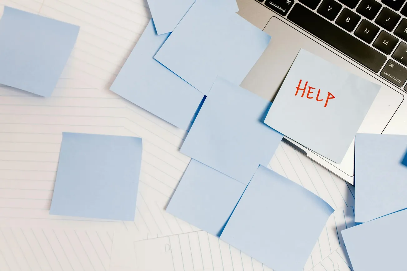 A cluttered workspace with blue sticky notes on a laptop, one showing "HELP" in red. Background features lined paper, conveying urgency and disorganization.