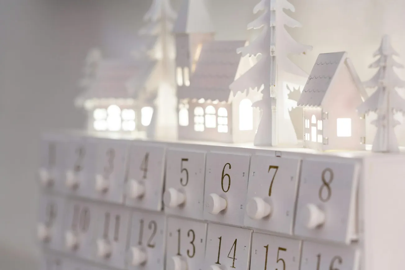 A white Advent calendar with numbered drawers and small illuminated houses and trees on top, evoking a serene, festive winter village scene.