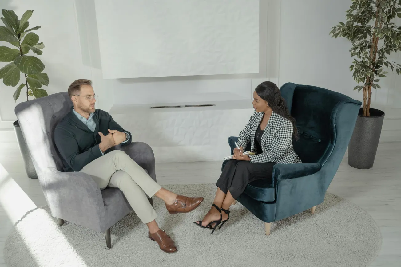 A man and woman sitting in armchairs engage in conversation in a bright, modern room with plants. The atmosphere is calm and professional.