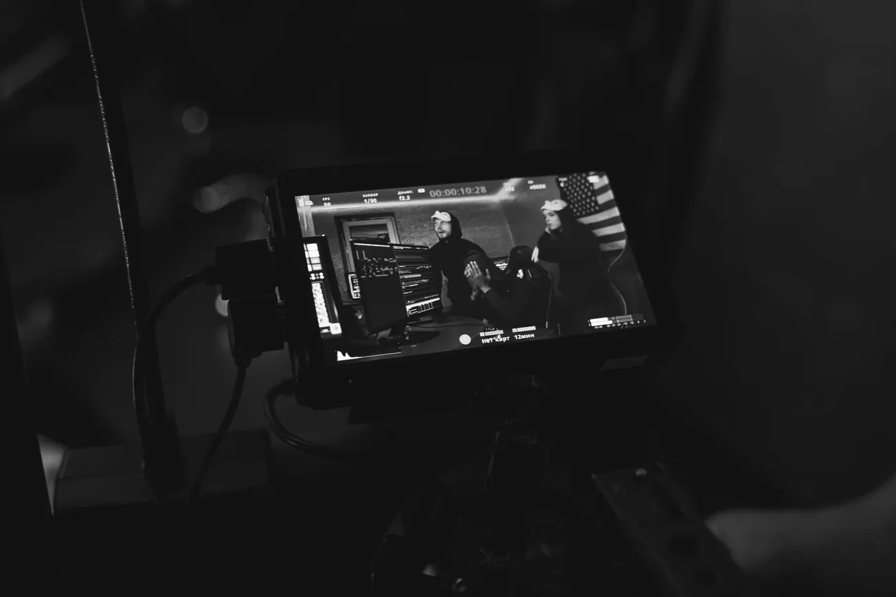 Black and white shot of a video camera screen showing three people in a room, possibly filming. An American flag is visible in the background.