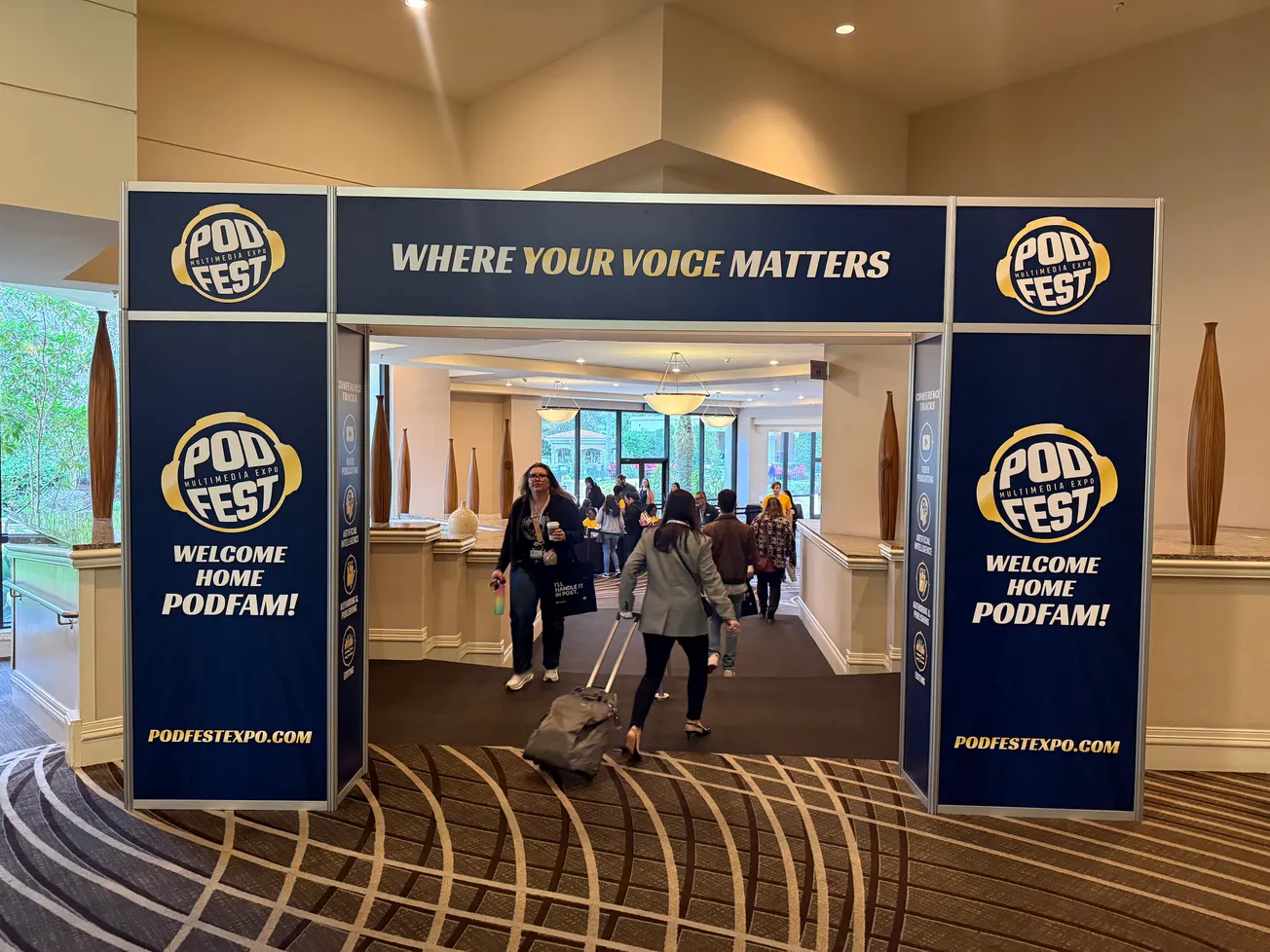 Entrance to PodFest conference with banners reading "Welcome Home PodFam!" Attendees with luggage enter a bright lobby, conveying excitement and community.