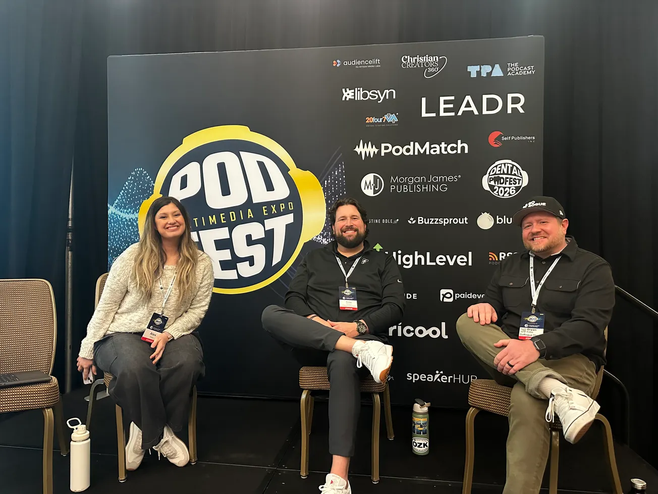 Three people seated in front of a "Podfest Multimedia Expo" backdrop. They are smiling and wearing conference badges. The mood is friendly and relaxed.