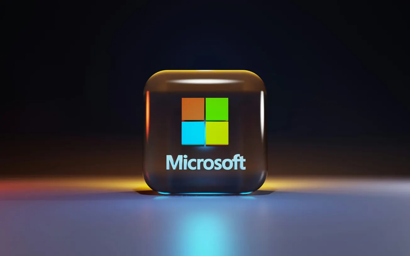 A glossy cube displays the Microsoft logo, with four colored squares above the word "Microsoft." Set against a dark background, the image conveys a sleek, modern tech feel.