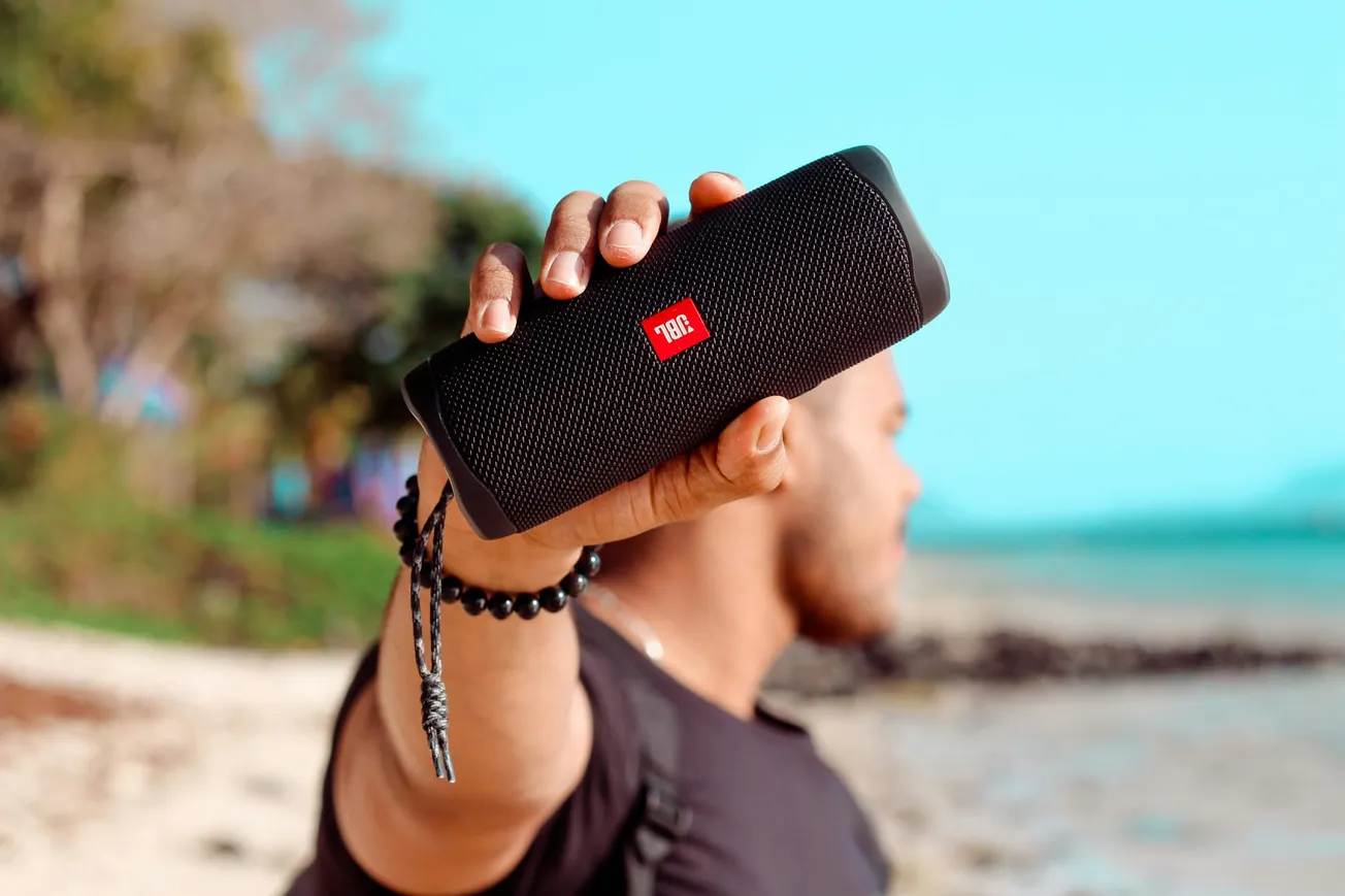 A person holds a black portable speaker at arm's length on a sunny beach. The focus is on the speaker, with blurred sand and trees in the background.
