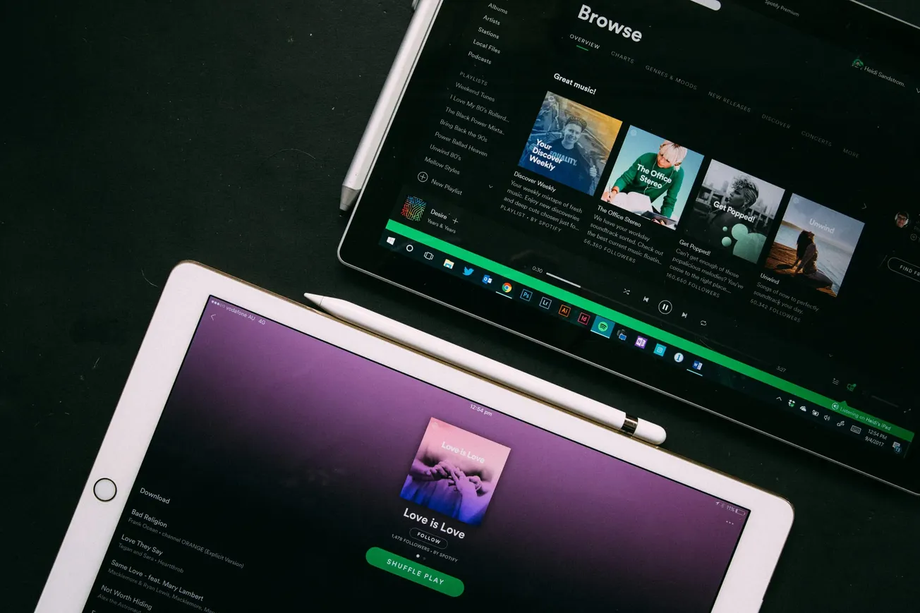 Tablets displaying Spotify apps on a dark surface. One shows a playlist with "Love is Love," the other shows a browse menu. Pens are nearby. Mood: Tech-focused.