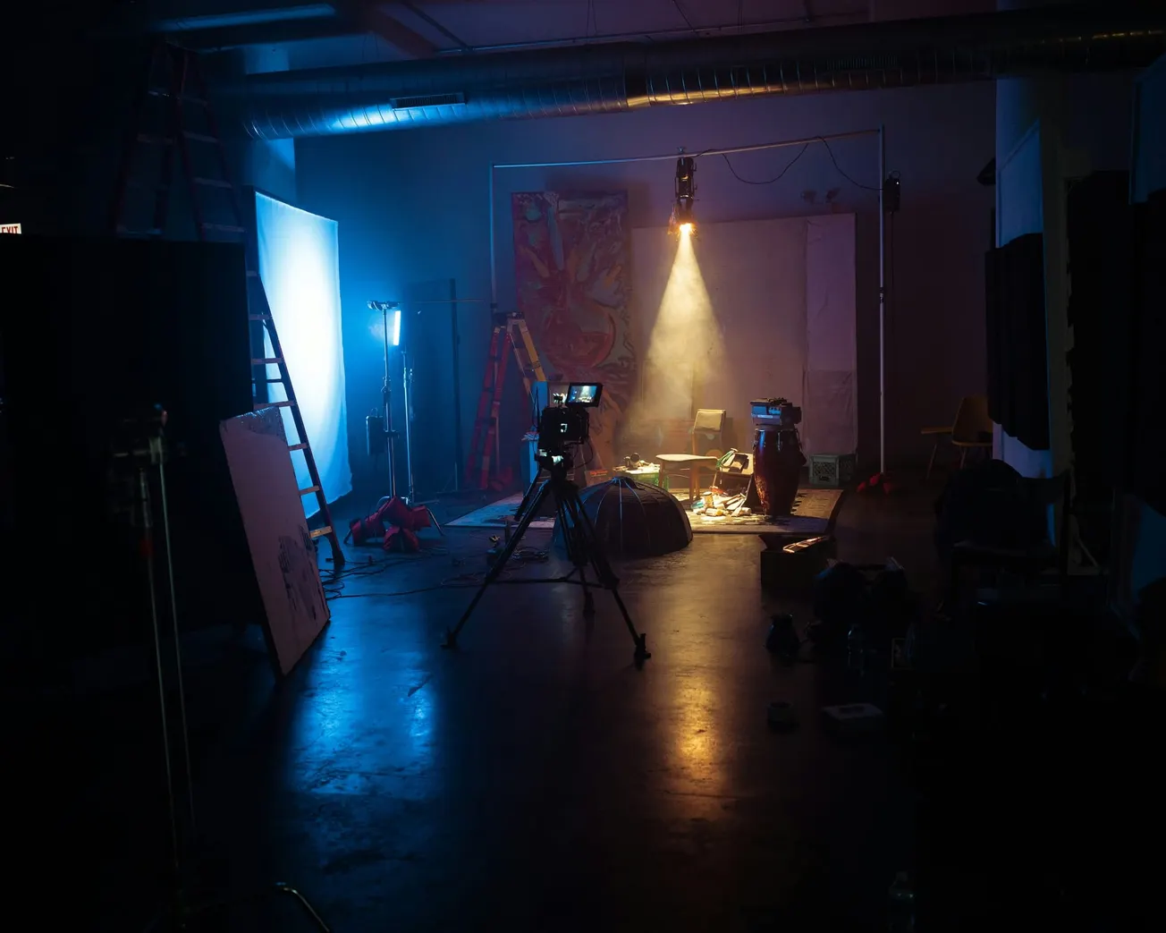 Dimly lit film set featuring tripods, cameras, and lighting equipment in a workshop-like studio. Ambient light casts a mysterious, creative mood.