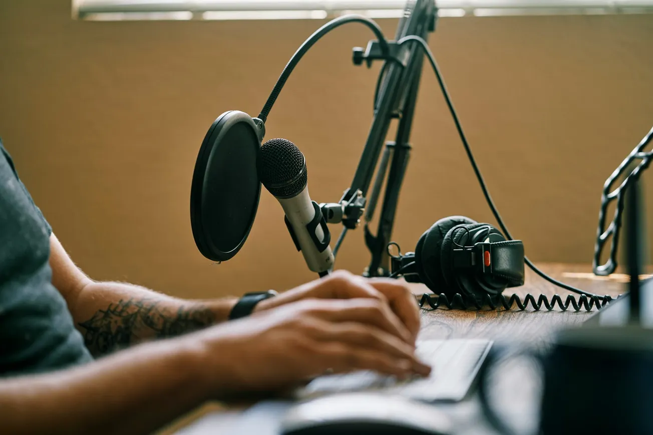 A close-up of a person typing with a tattooed arm at a wooden desk. A professional microphone, pop filter, and headphones are nearby, suggesting a podcast setup.