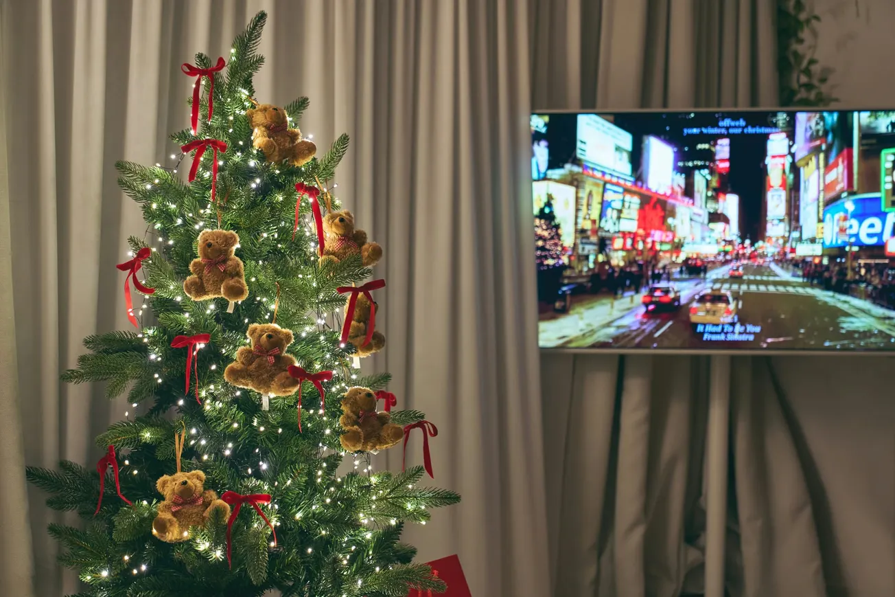 A decorated Christmas tree with teddy bear ornaments and red ribbons is lit with white lights. A TV displays a brightly-lit cityscape at night.
