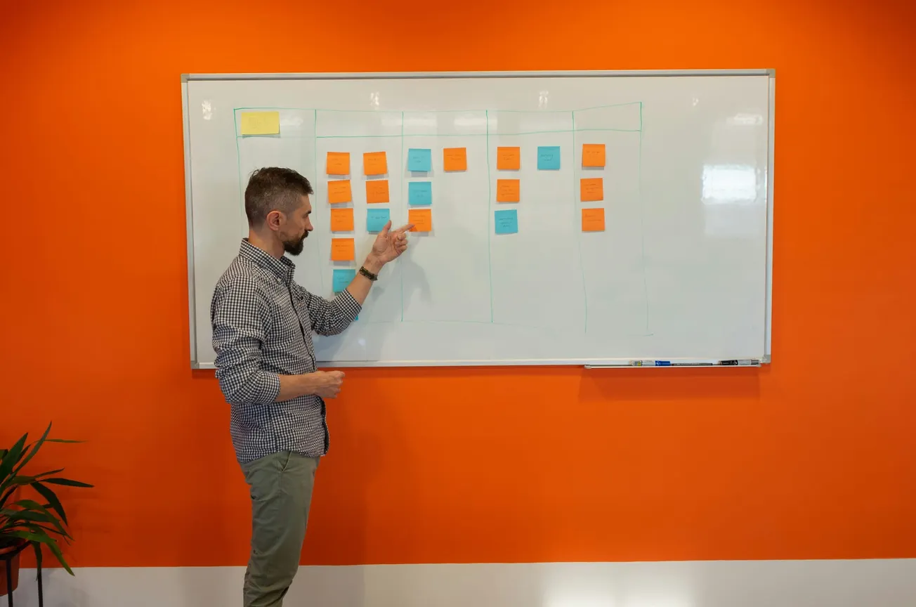 A man stands in front of a whiteboard covered with colorful sticky notes, pointing as if explaining something. The room has a vibrant orange wall, conveying a lively atmosphere.