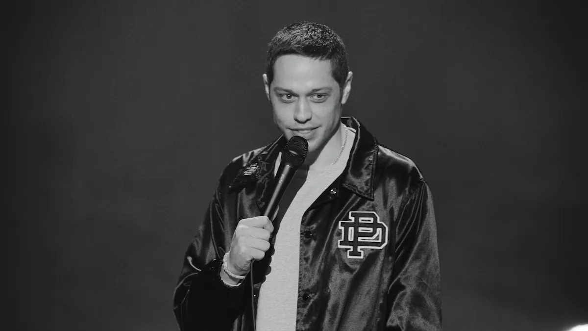 Pete Davidson wearing a black satin jacket holds a microphone, smiling slightly, with a neutral background. The scene conveys a casual, comedic tone.