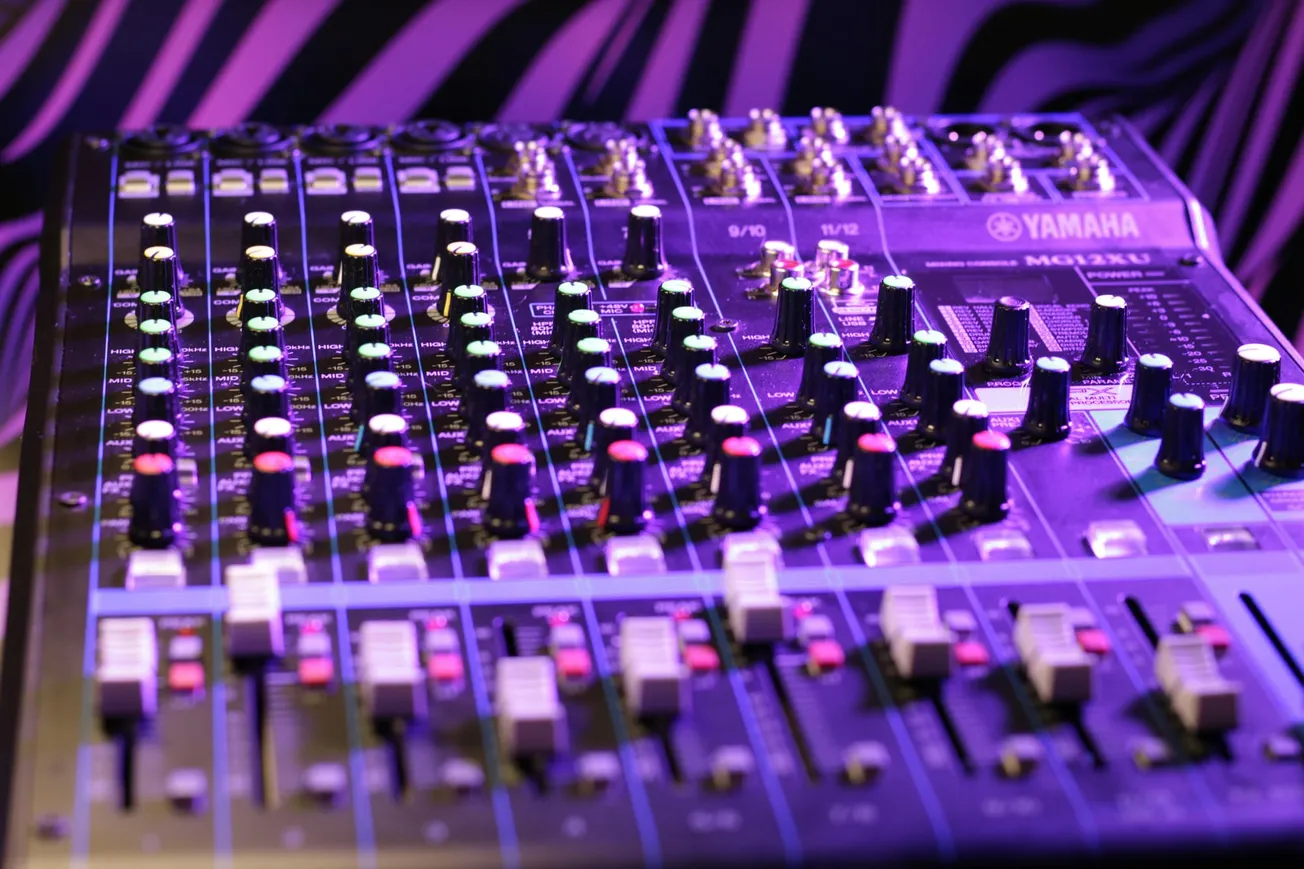 Close-up of a Yamaha MG32/14FX audio mixer with numerous knobs and sliders, lit by purple and blue lights, creating a vibrant, technical ambiance.