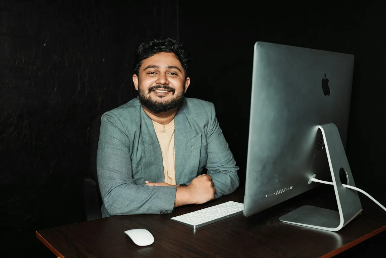 A person with a beard smiles warmly at a desk with a large computer, keyboard, and mouse against a dark background, creating a professional yet relaxed atmosphere.