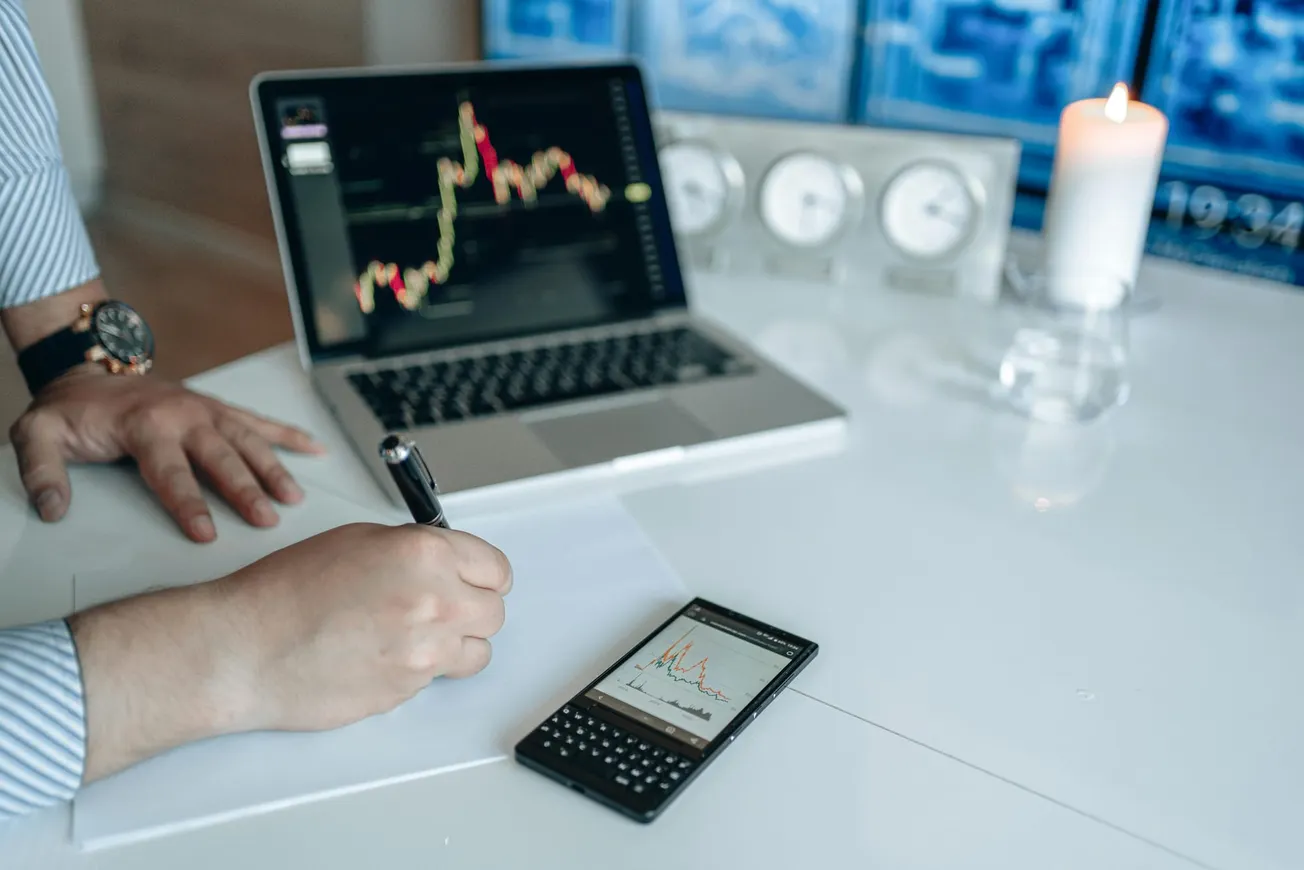A person writes on paper at a desk, with a laptop showing stock charts and a smartphone displaying graphs. A lit candle adds a calm atmosphere.