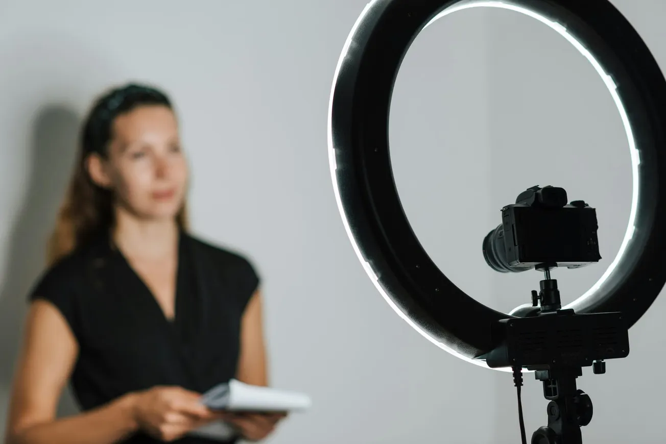 A woman stands blurred in the background holding papers, while a camera is focused in the foreground with a bright ring light, creating a professional setup.
