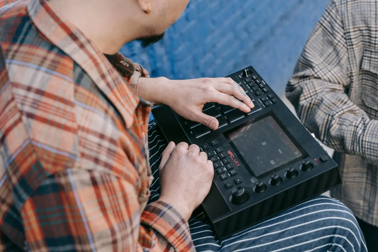 Person in plaid shirt operates a music production device outdoors, fingers pressing buttons on a digital pad, conveying a focused and creative mood.