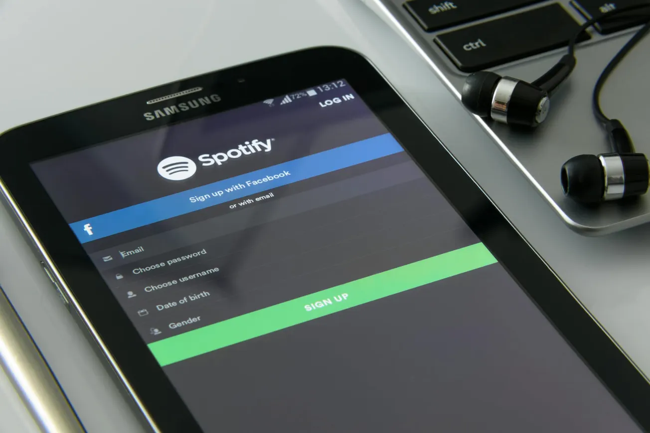 A tablet displays the Spotify sign-up screen, offering options via Facebook or email. Nearby are earbuds and a laptop keyboard, conveying a tech-savvy mood.
