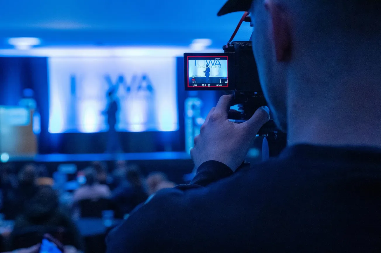 A cameraman films a speaker on stage at a conference. The scene is lit in blue, and the camera screen captures the speaker in focus.