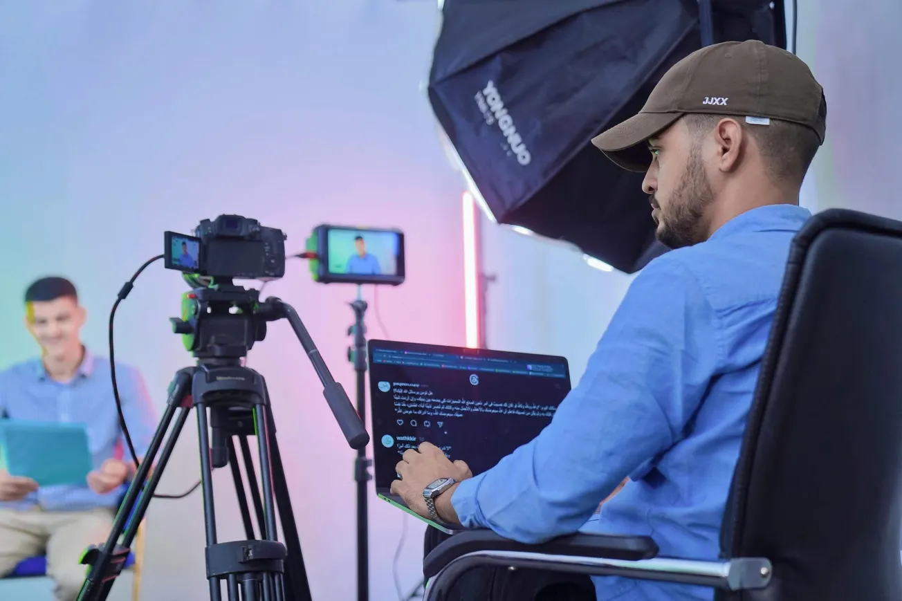 Man in blue shirt and cap, seated in a studio, uses a laptop while filming a person on camera. Soft lighting and focused, professional setup.