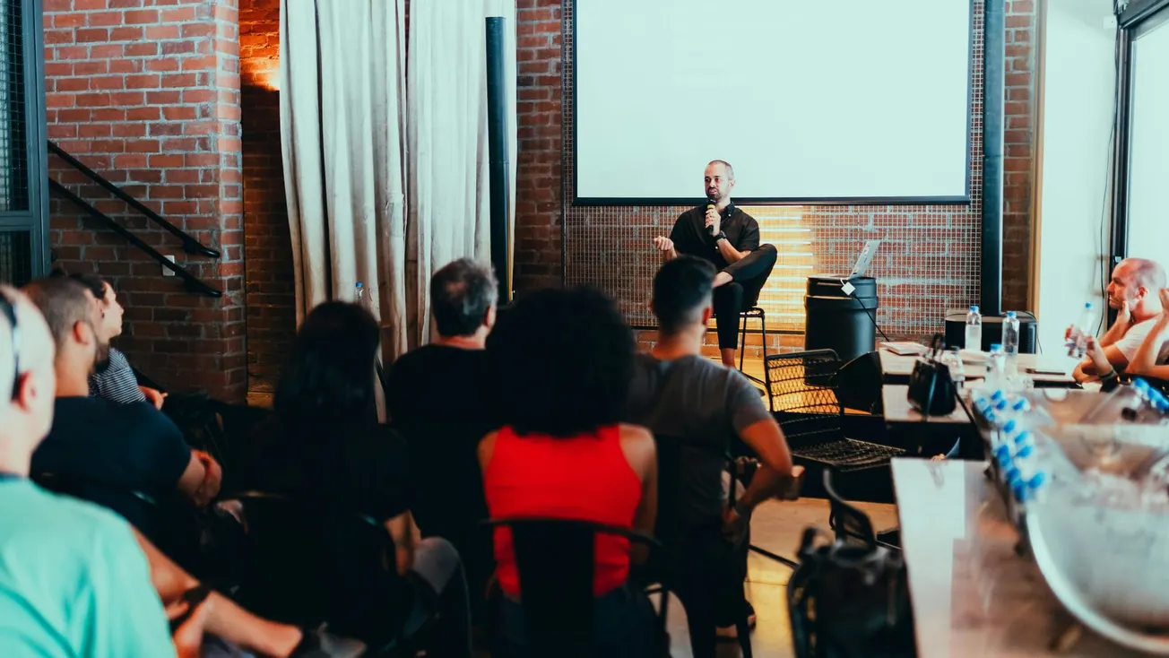 A speaker sits on a stool in front of an audience in a cozy, brick-walled room. The atmosphere is attentive, with viewers focused on the presentation.