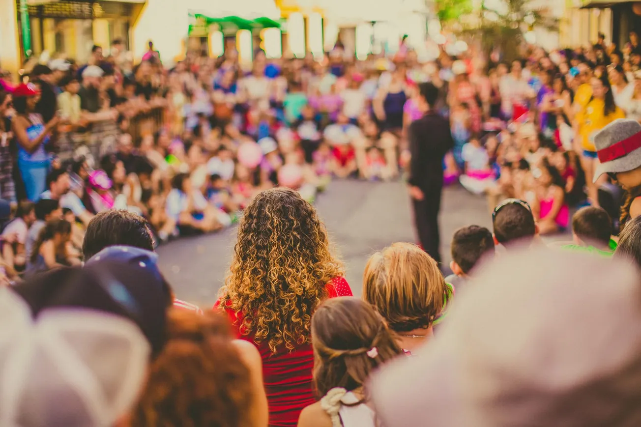 A large crowd gathers outdoors around a street performer. The scene is lively and colorful, with people sitting and standing, enjoying the entertainment.