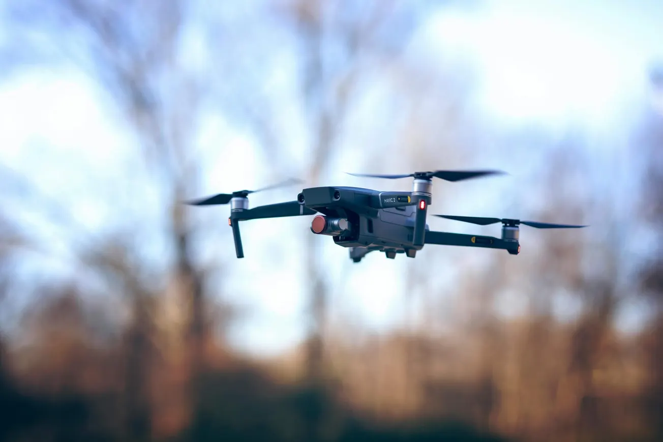 A black drone with four propellers hovers mid-air against a blurred background of bare trees. The sky is clear, conveying a serene outdoor setting.