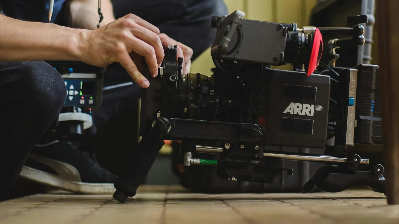 Hands adjust a professional ARRI camera on a movie set. The floor is tiled, and the scene conveys a focused, technical atmosphere.