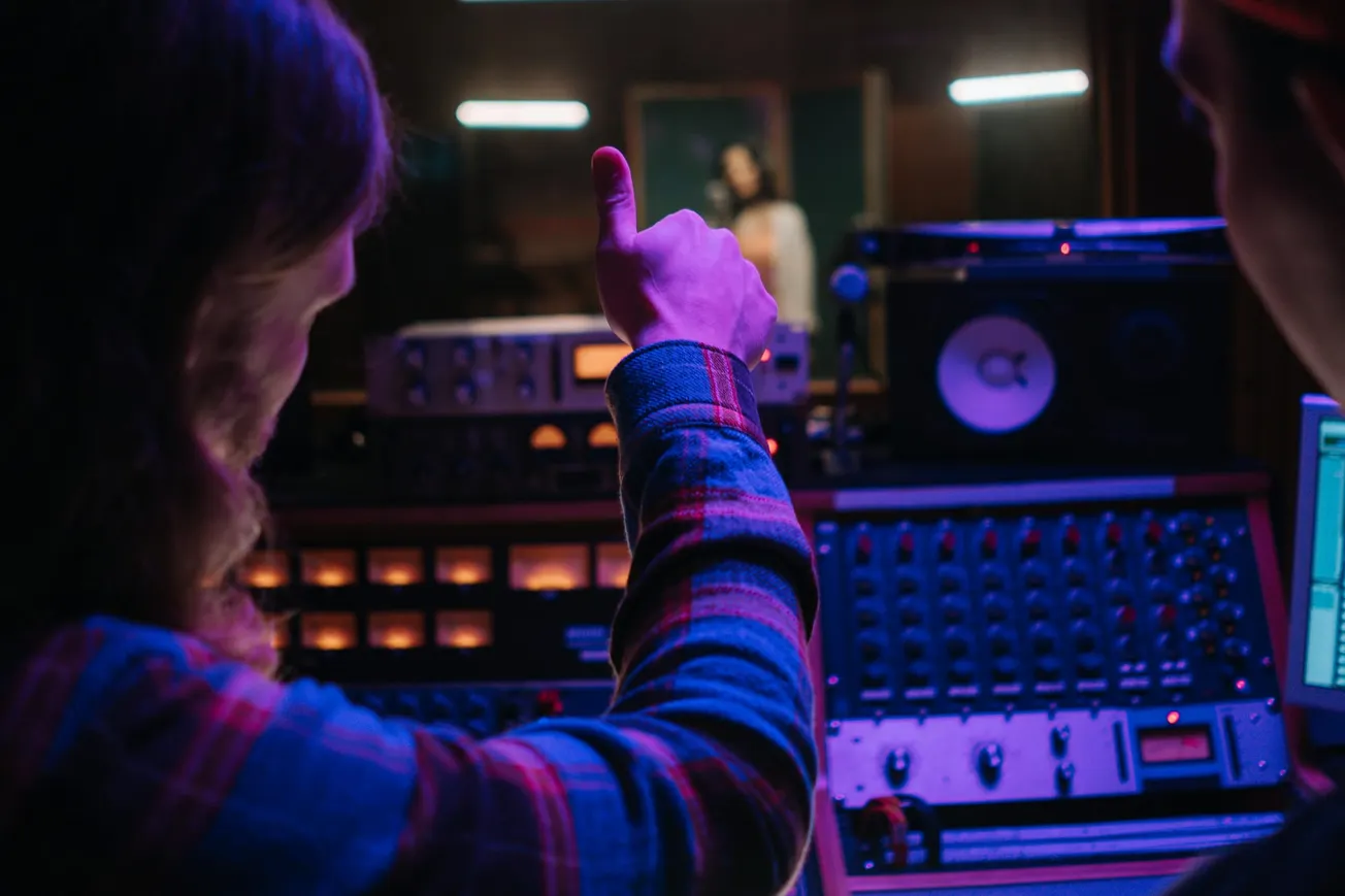 A person gives a thumbs-up in a dimly lit recording studio, surrounded by audio equipment. Another person sings in the booth, creating a focused atmosphere.