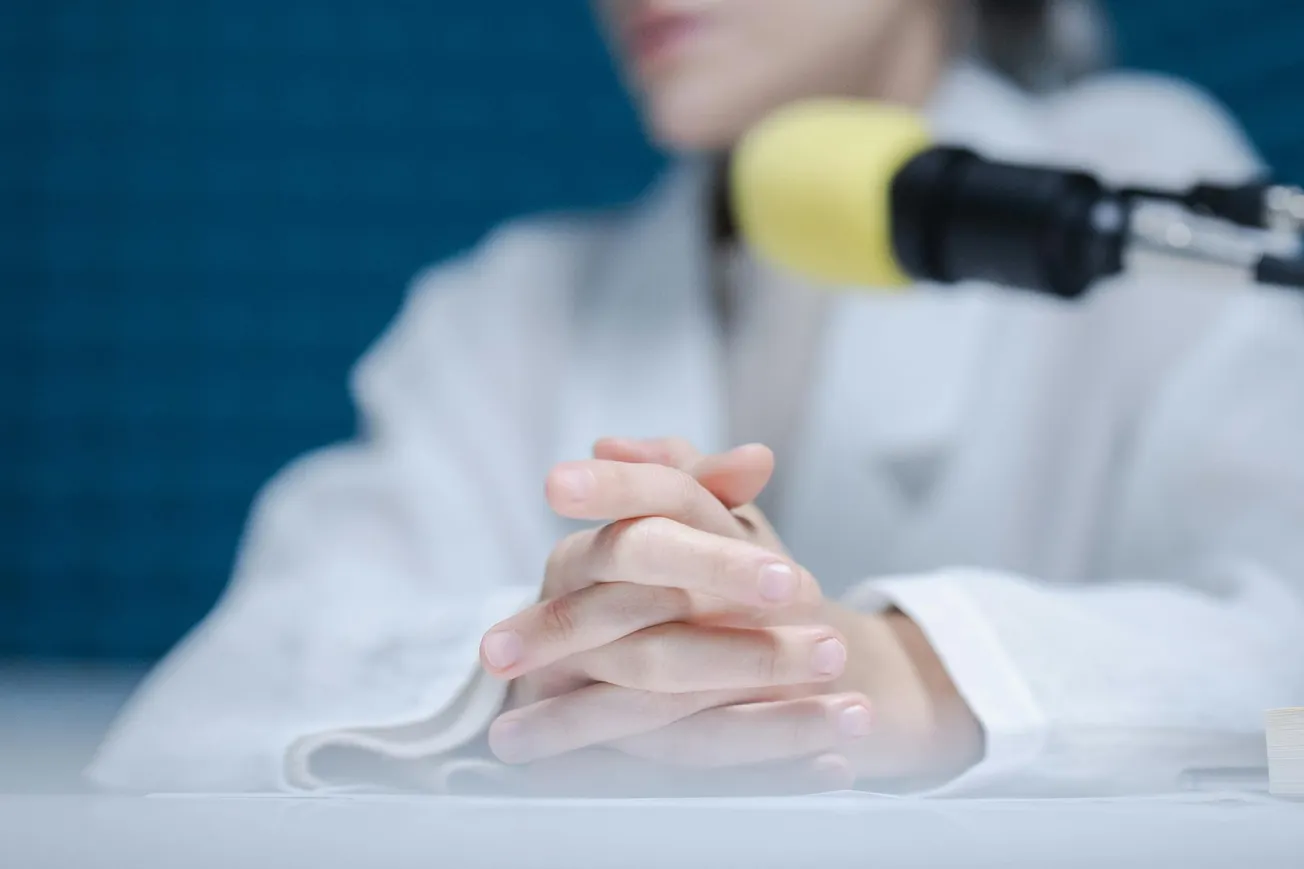 A person in a white coat sits at a table, hands clasped, with a microphone nearby. The setting conveys a serious or professional tone.