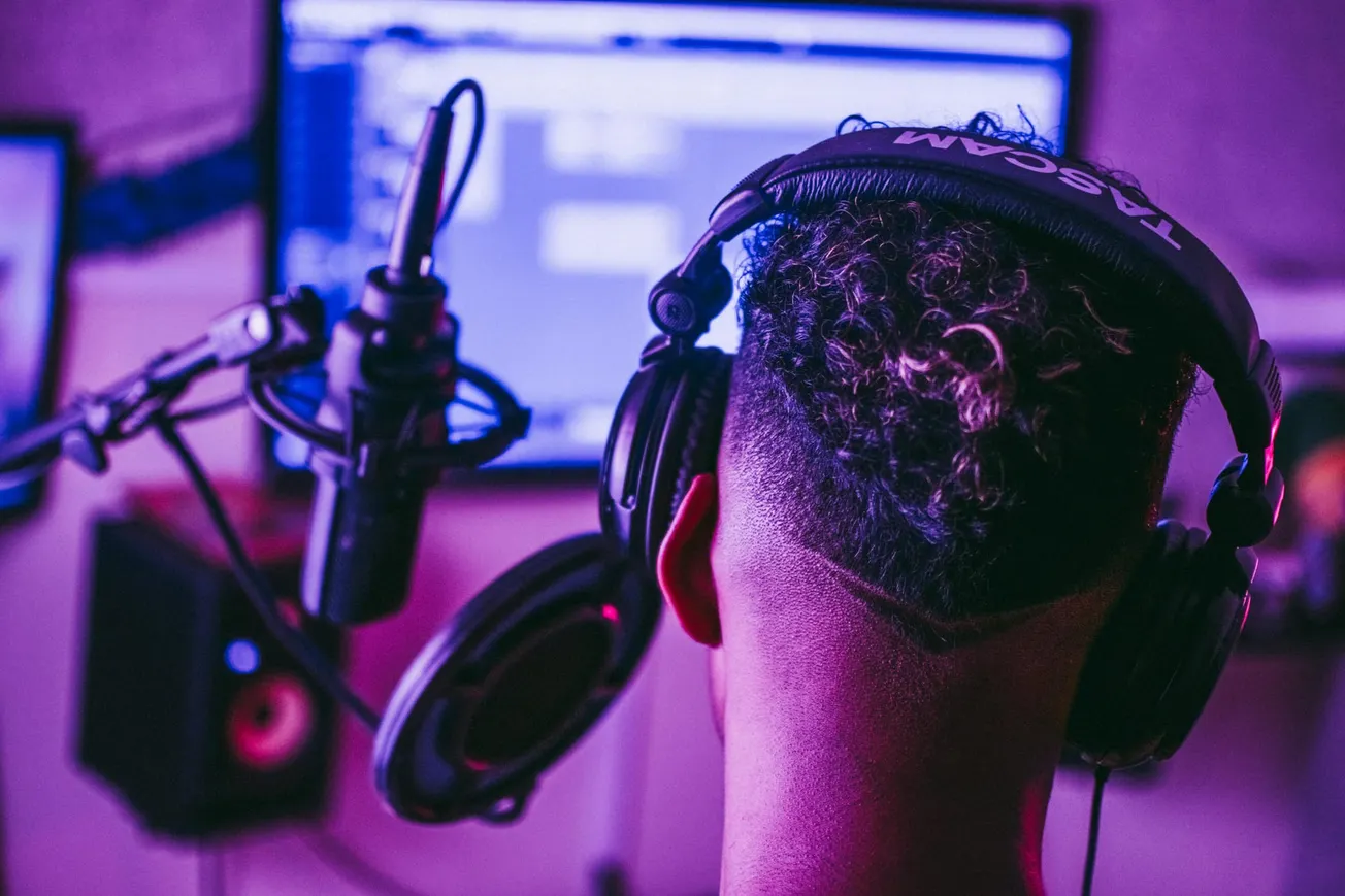 A person wearing large headphones sits in front of a microphone, facing a computer screen. The room has a soft purple glow, creating a creative ambiance.