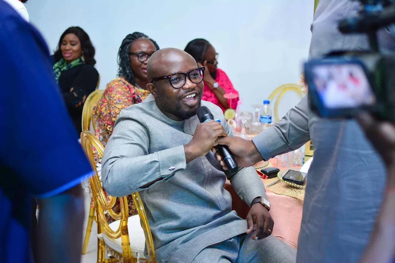 A smiling man in glasses and grey outfit speaks into a microphone while seated at a lively event. Others in colorful attire converse in the background.
