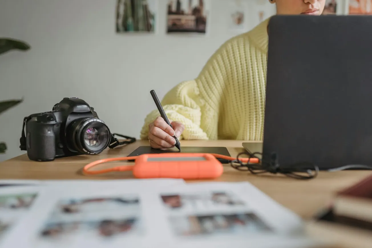 Person in a yellow sweater using a graphics tablet at a desk with a camera and hard drive. Photos are blurred in the foreground and background.