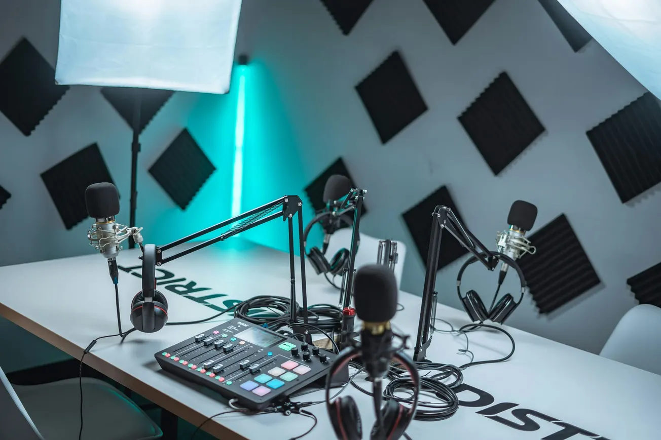 Podcast studio with a white table, equipped with microphones on adjustable arms, a mixer, and headphones. Soundproof foam panels on walls; a calm, focused setting.