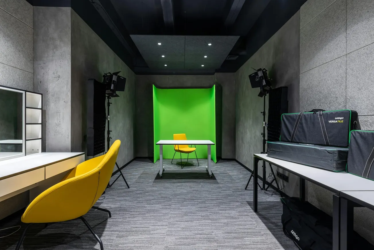 A modern studio with green screen backdrop, yellow chairs, a desk, and professional lighting. The room is sleek with gray walls and carpet.