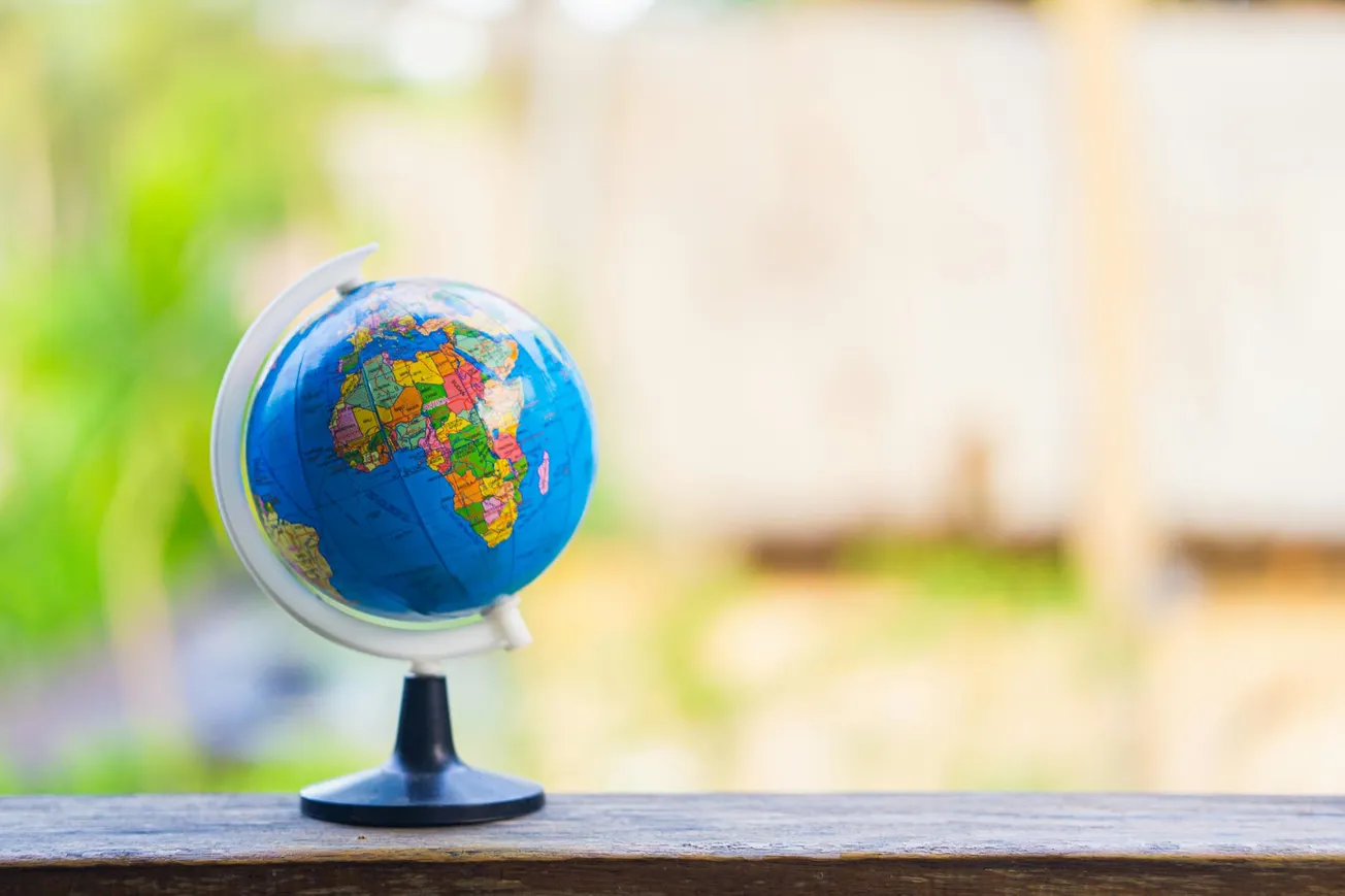 A small globe displaying Africa sits on a wooden ledge, with a blurred, sunny outdoor background. The scene is bright and colorful, evoking exploration.