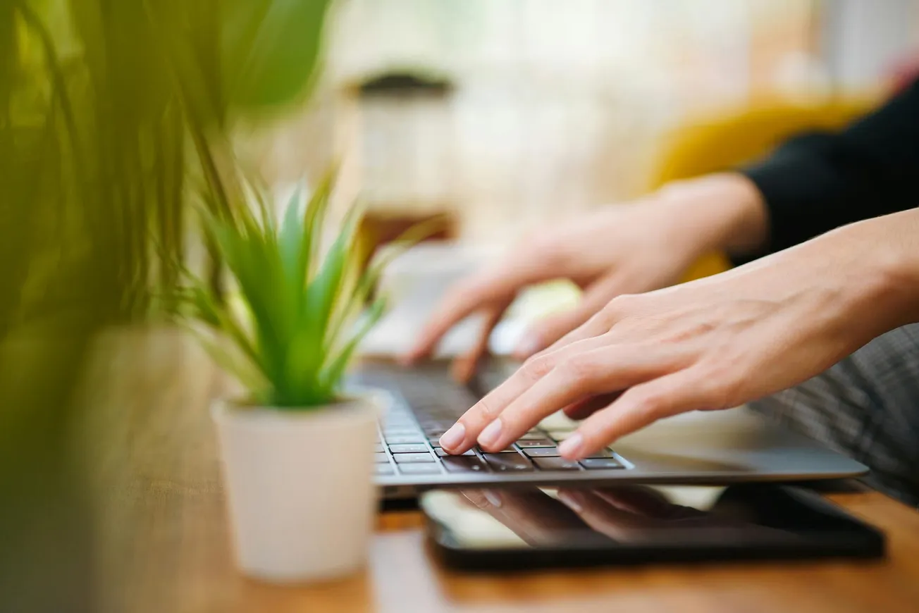 Hands typing on a laptop keyboard on a wooden desk. A smartphone and a small potted plant sit nearby, with a blurred, bright background.