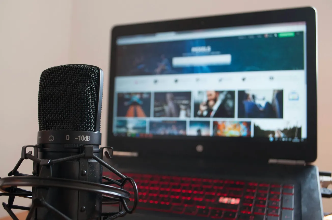 Close-up of a microphone with a laptop in the background. The laptop screen shows a blurred website interface. The scene suggests a podcast or streaming setup.