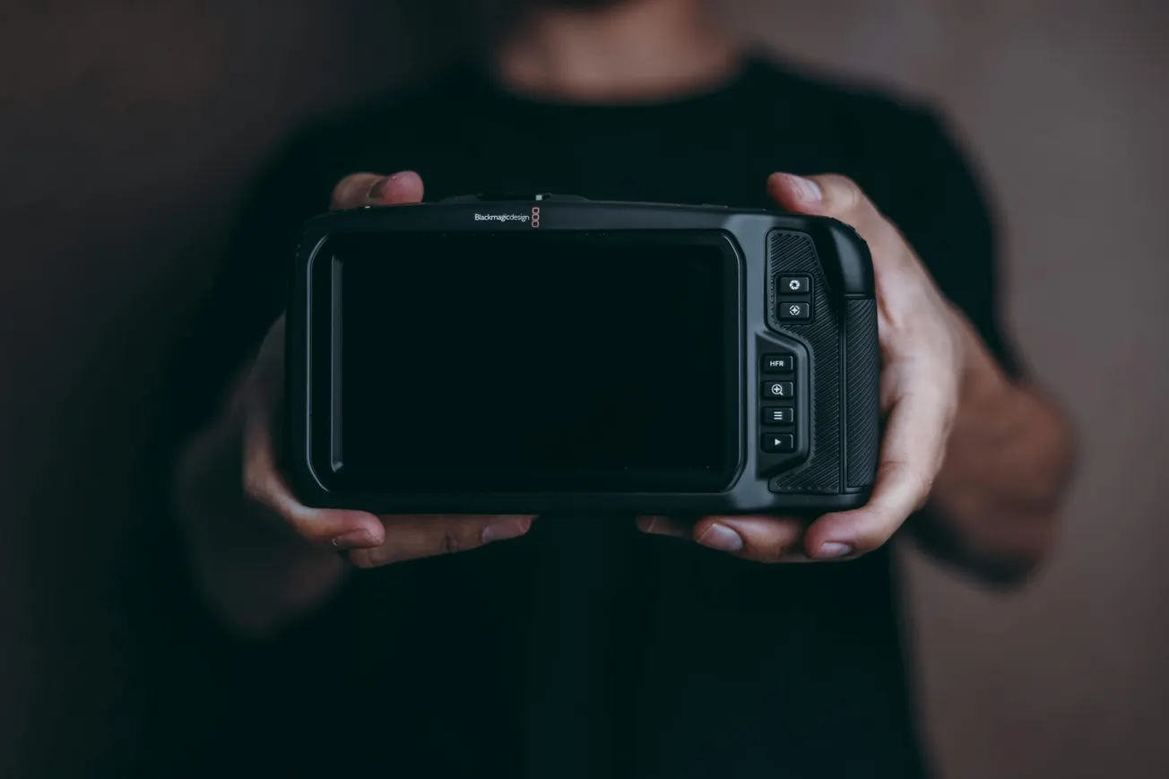 Person holding a large, black camera with a screen facing forward, against a dark background. The mood is focused and professional.