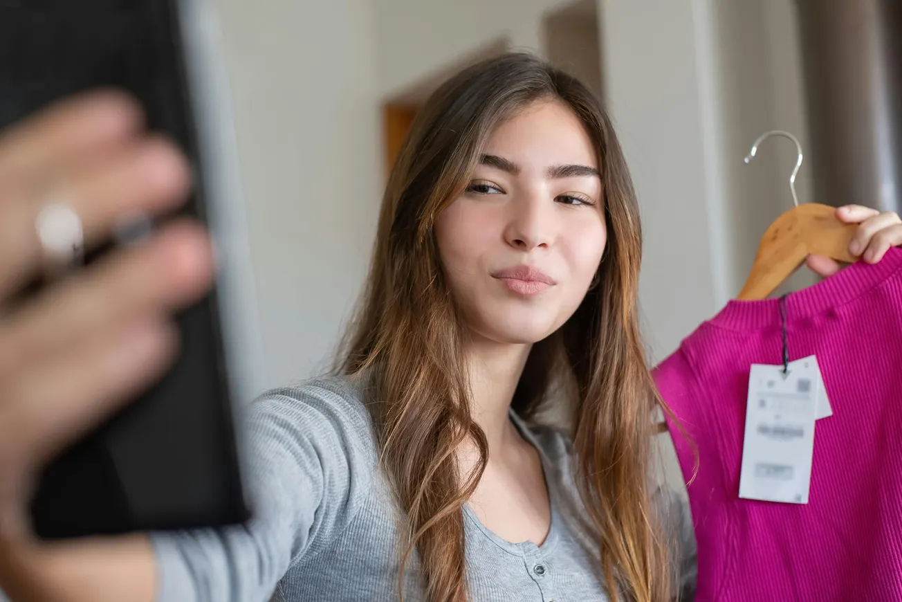 Young woman with long brown hair takes a selfie while holding a bright pink top on a hanger, smiling slightly, in a modern indoor setting.
