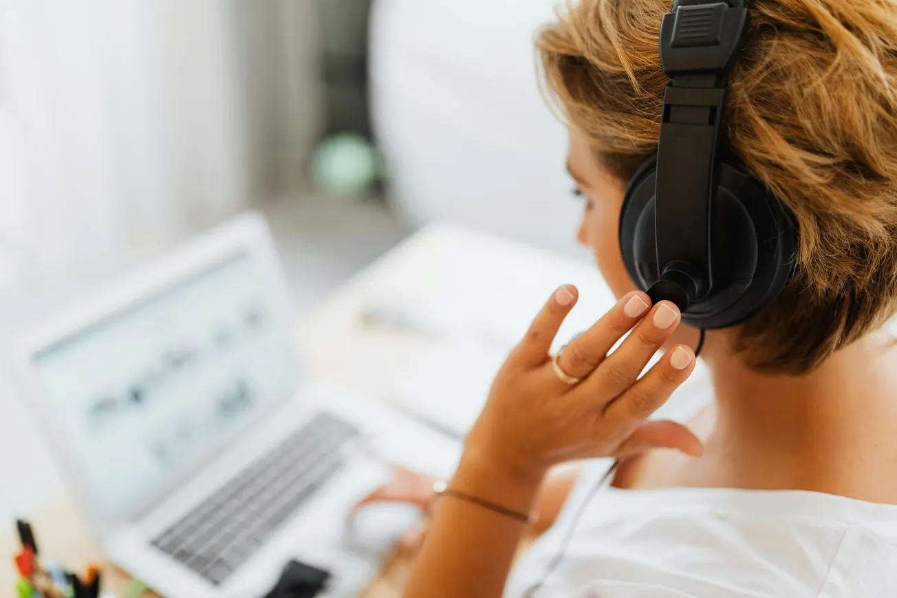 A person wearing headphones is seen from behind, seated at a desk with a laptop. They are focused and engaged, suggesting a work or study setting.