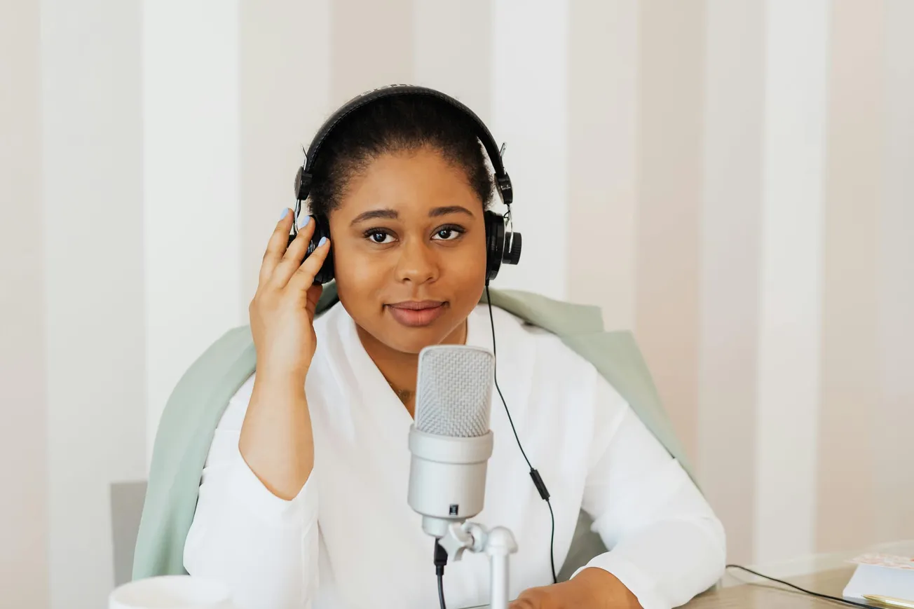 Woman in white blouse wearing headphones, sitting at a desk with a microphone. She looks towards the camera with a calm expression.