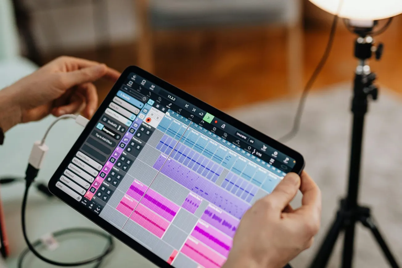 Hands hold a tablet displaying a colorful music production app with various tracks and controls. The scene conveys creativity and technology.