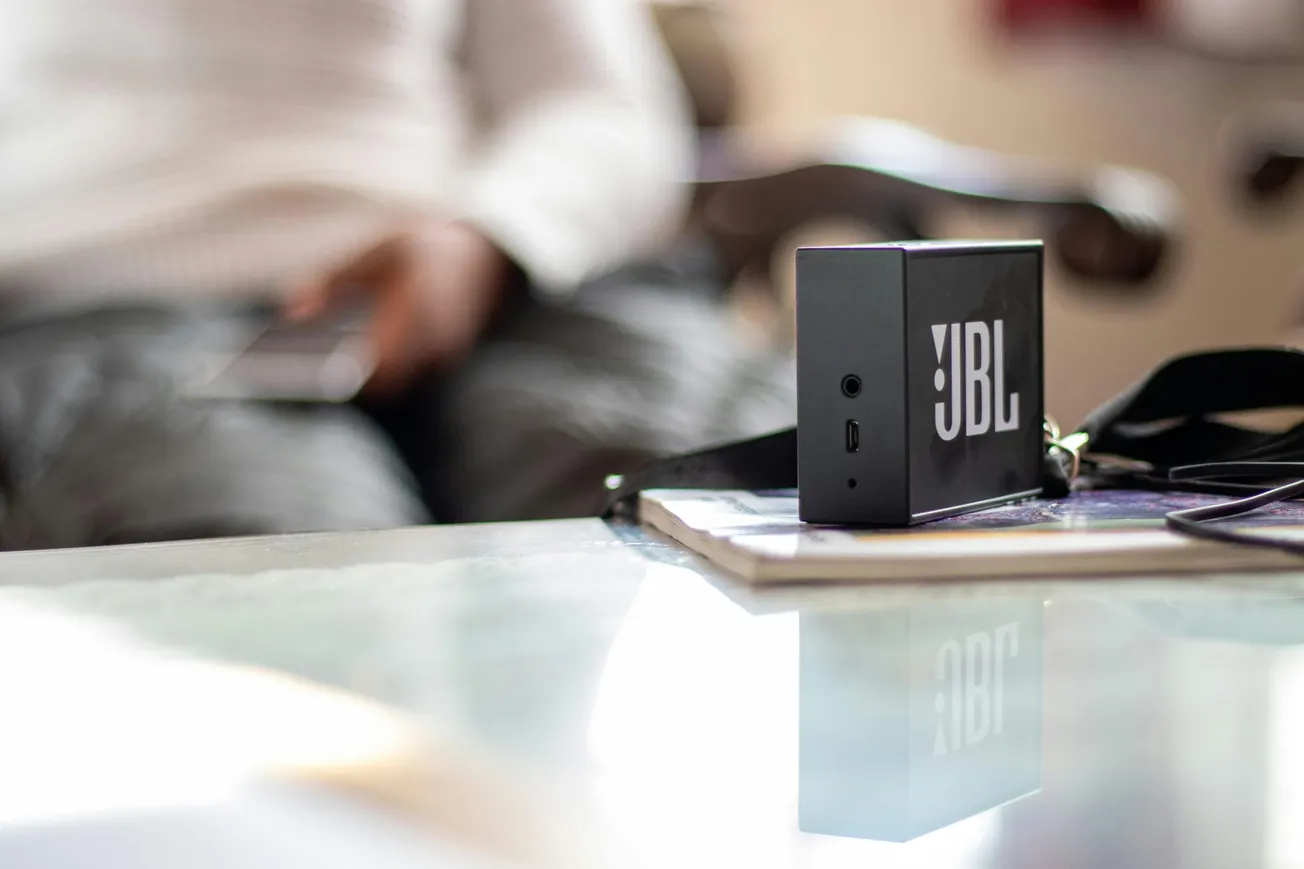 A black JBL Bluetooth speaker rests on a table beside closed books, slightly blurred background shows a person with a phone.