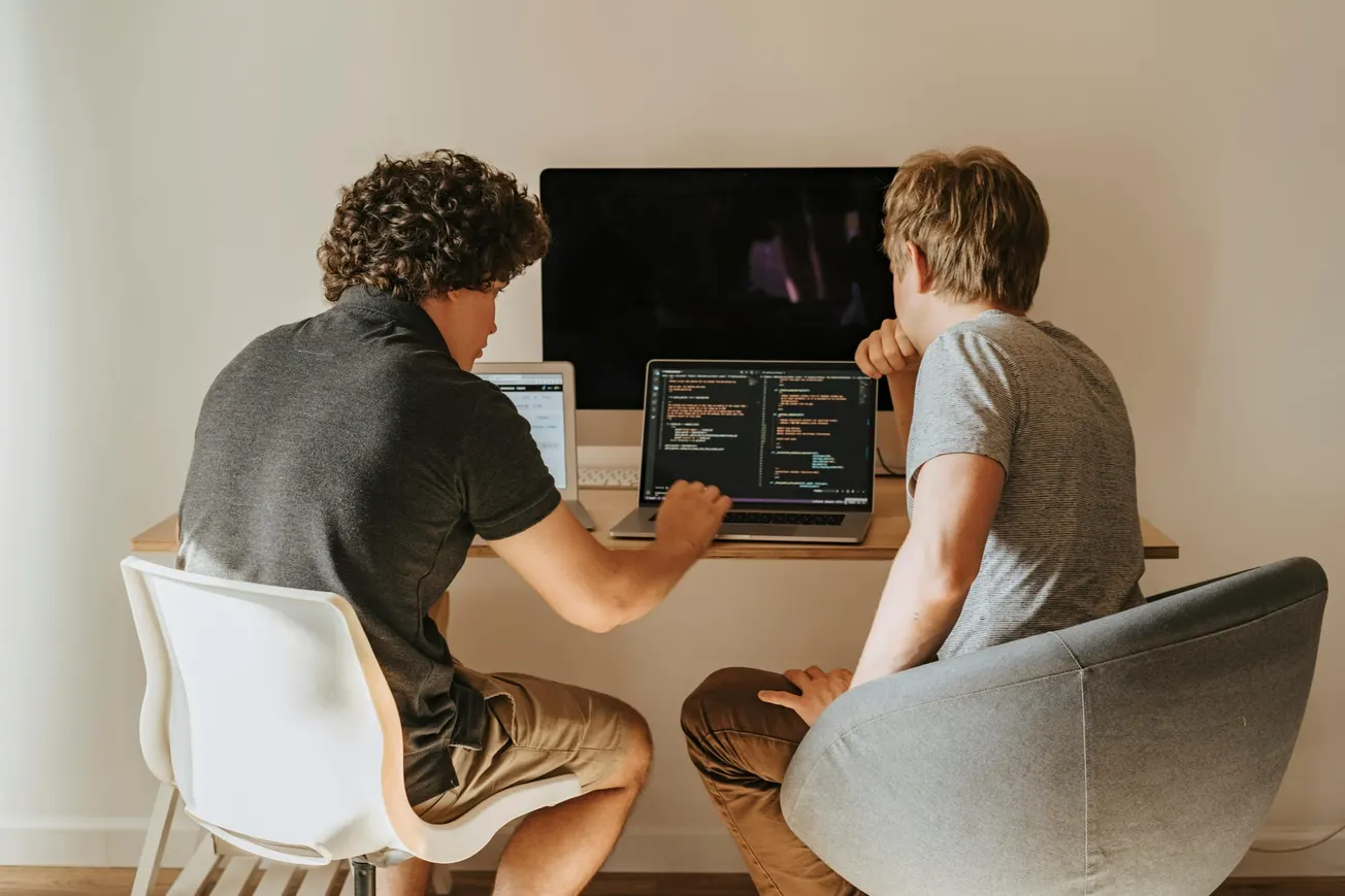 Two people sit at a desk collaborating on laptops displaying code. A large monitor is in the background. The atmosphere is focused and collaborative.
