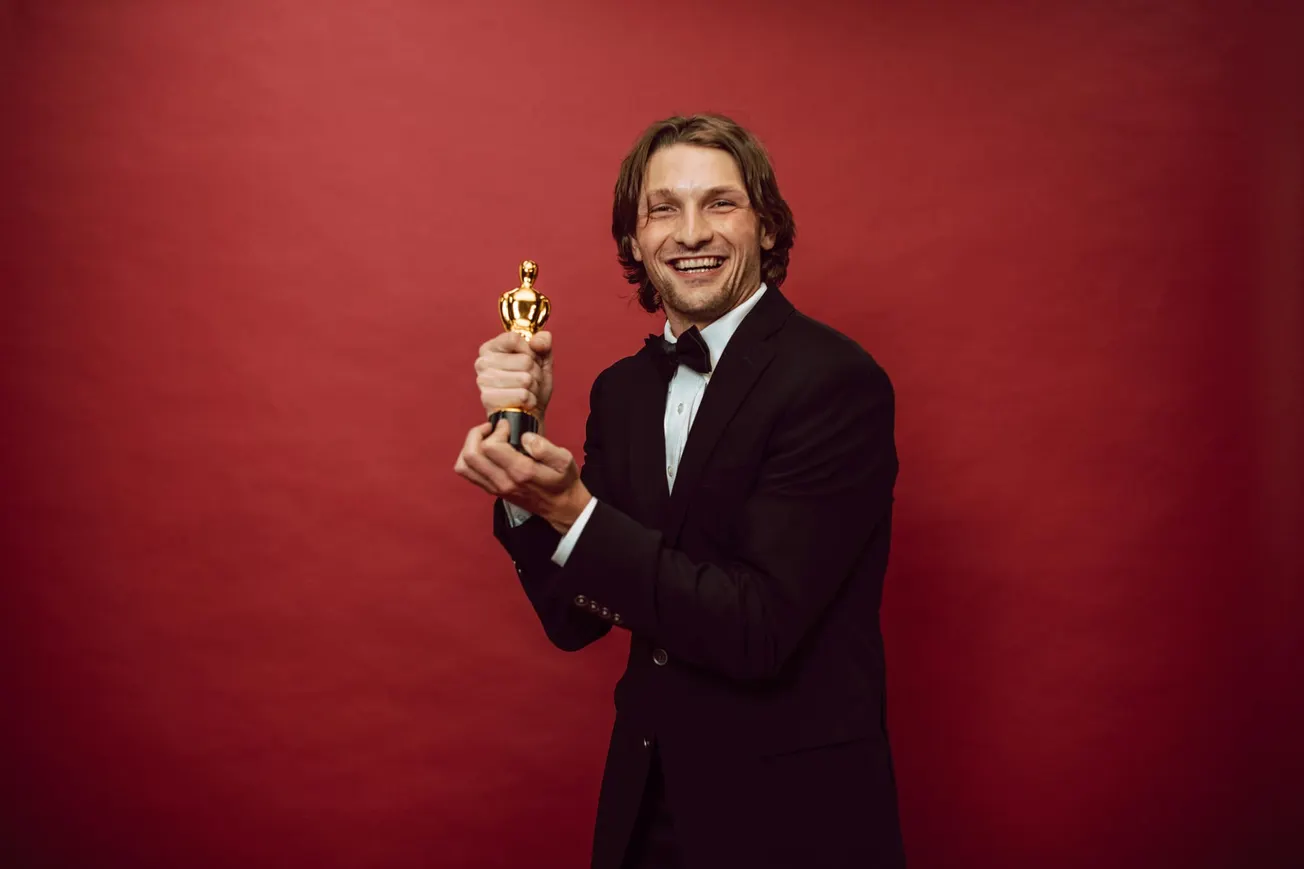 A smiling man in a black tuxedo holds a gold trophy against a red background, conveying joy and celebration in a formal setting.
