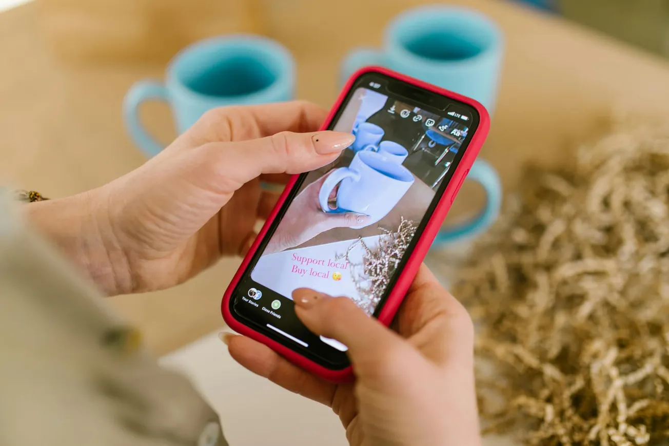 A person holds a smartphone showing a photo of blue mugs with the text "Support local, Buy local" beside similar mugs on a table. Warm and inviting tone.