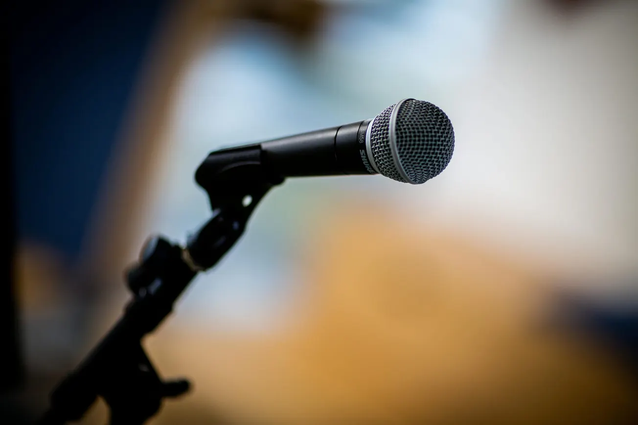 Close-up of a microphone on a stand, angled slightly upward, against a blurred background. The image conveys anticipation and focus.