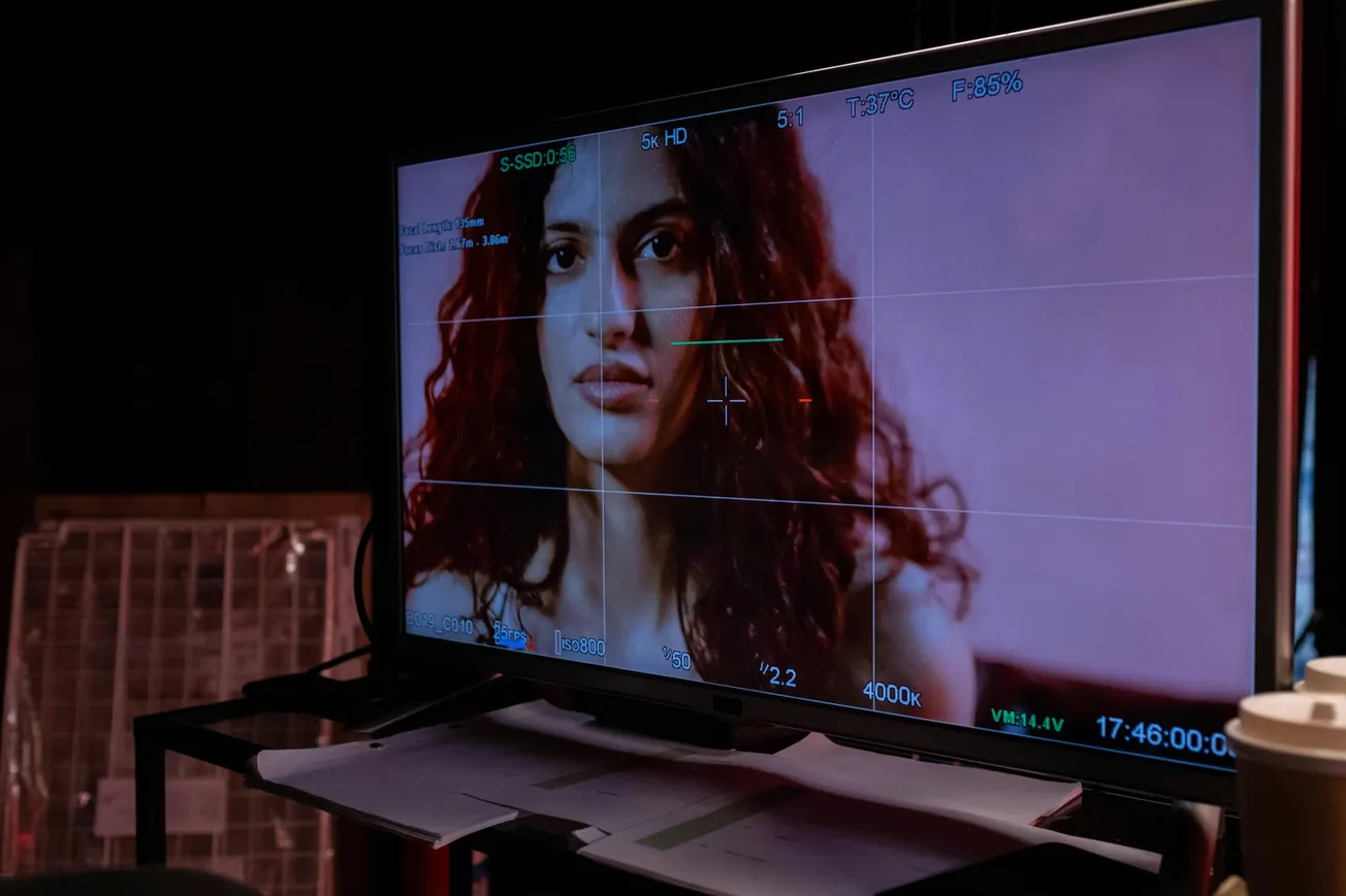 A monitor displays a close-up of a woman with curly hair, her expression serious. The screen shows camera settings. Scene feels focused and cinematic.