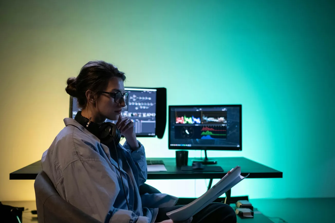 A person in glasses and headphones studies documents in a dimly lit room. Two monitors display graphs, casting a green and yellow glow, conveying focus.