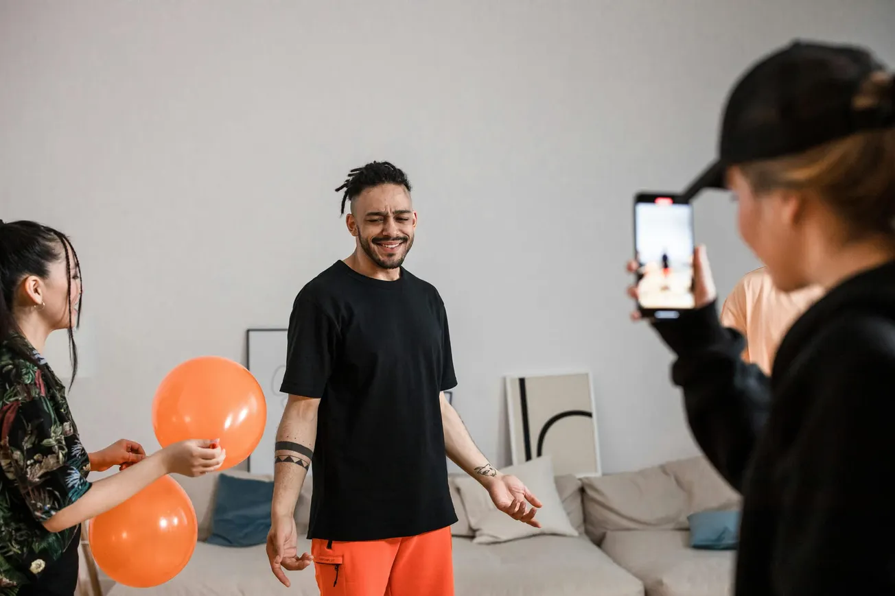 Three people in a living room: one holding orange balloons, another in orange pants smiling with eyes closed, and a third taking a photo with a phone.