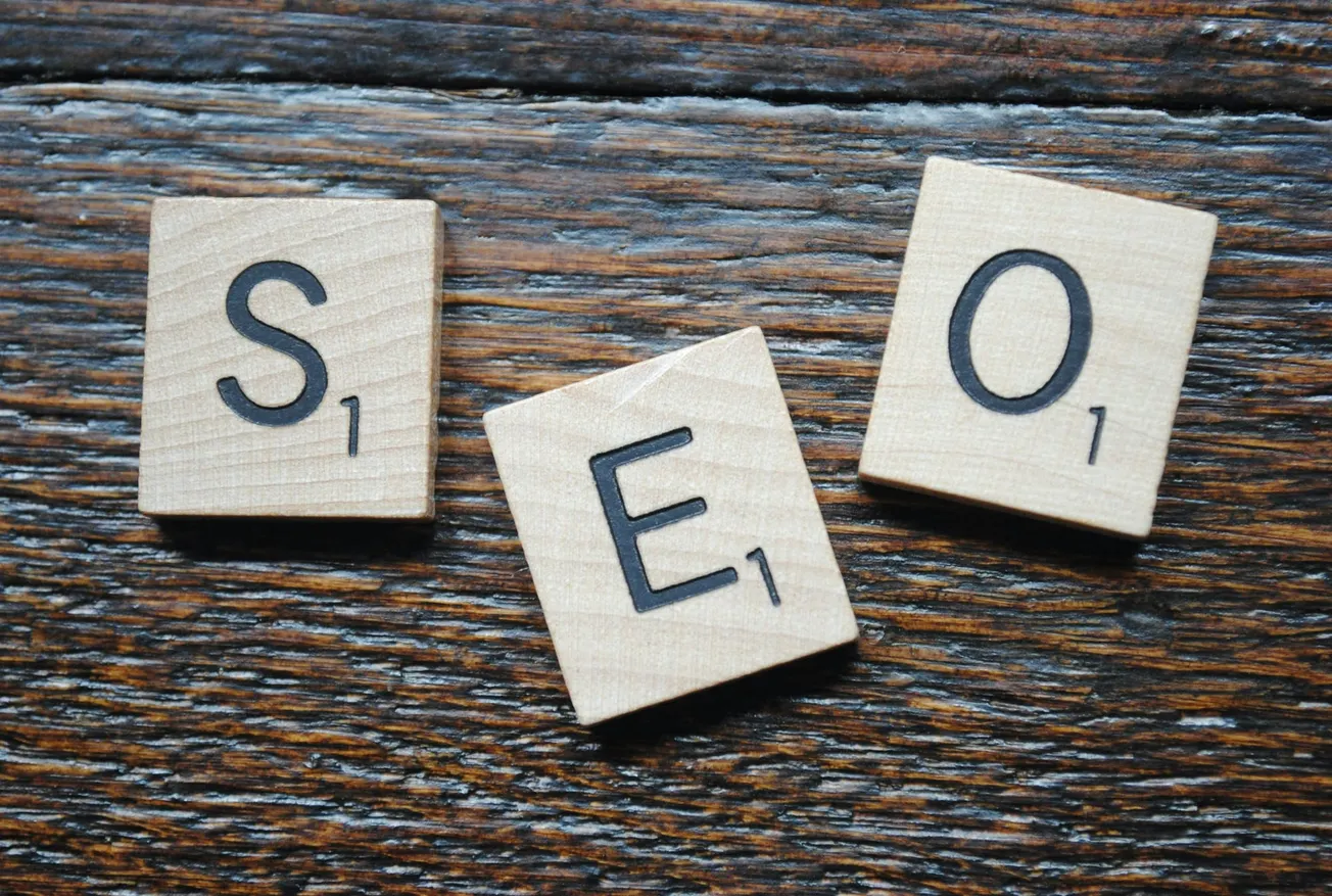 Scrabble tiles spelling "SEO" sit on a textured wooden surface. Each tile has a letter and scoring number, conveying a casual, strategic theme.